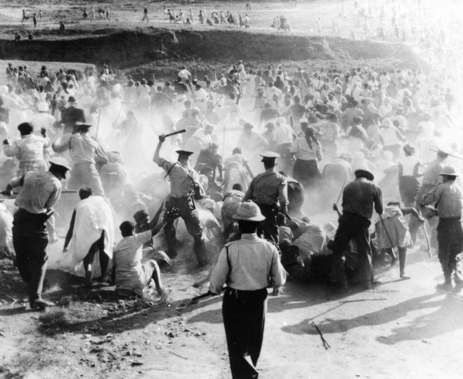 Black and white photo of a chaotic scene. A crowd of people is attacked by police officers with clubs. Some people are standing, some are sitting or lying on the ground. Some are running away. Smoke /or dust adds to the confusion of the moment.